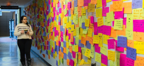 A student walking through a hallway looking at a wall covered in colourful post it notes