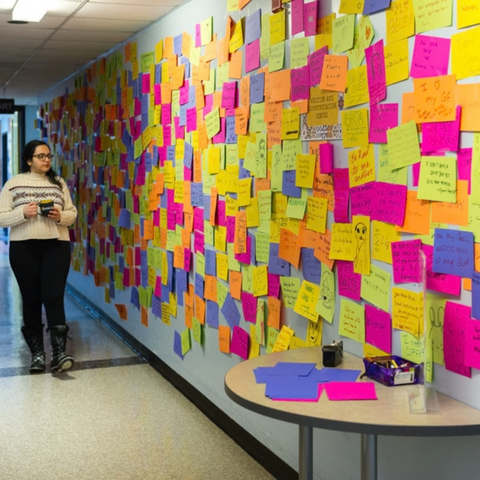 A student walking through a hallway looking at a wall covered in colourful post it notes