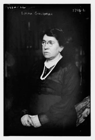 A black and white photo of a middle aged white woman, Emma Goldman, with short hair, wearing glasses, pearls, and a black dress, posed seated and looking to the left with folded hands.