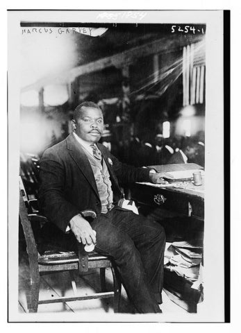 A black and white photo of a black man in a suit with cropped hair seated on a chair and posed facing to the right.
