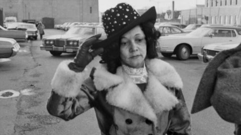 A black and white image of a black woman in a coat holding onto her hat.