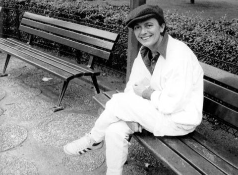 A black and white photo of a white woman smiling at the camera while sitting on a bench outdoors. She is wearing white trousers and a white collared shirt with a hat.