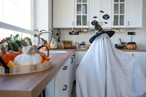  A child in sheets with cutout for eyes like a ghost costume in the kitchen decorated for the Halloween holiday. A kind little funny ghost. Halloween Party.