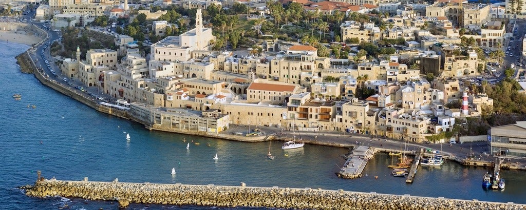 Aerial view of the port of Jaffa. There is a roadway all along the waterfront, soem sailboats in the water, and square, stone balding in the background