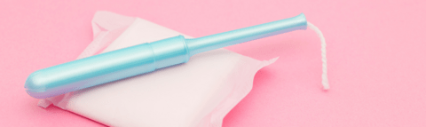 A blue tampon rested on top of a menstrual pad with a pink background.
