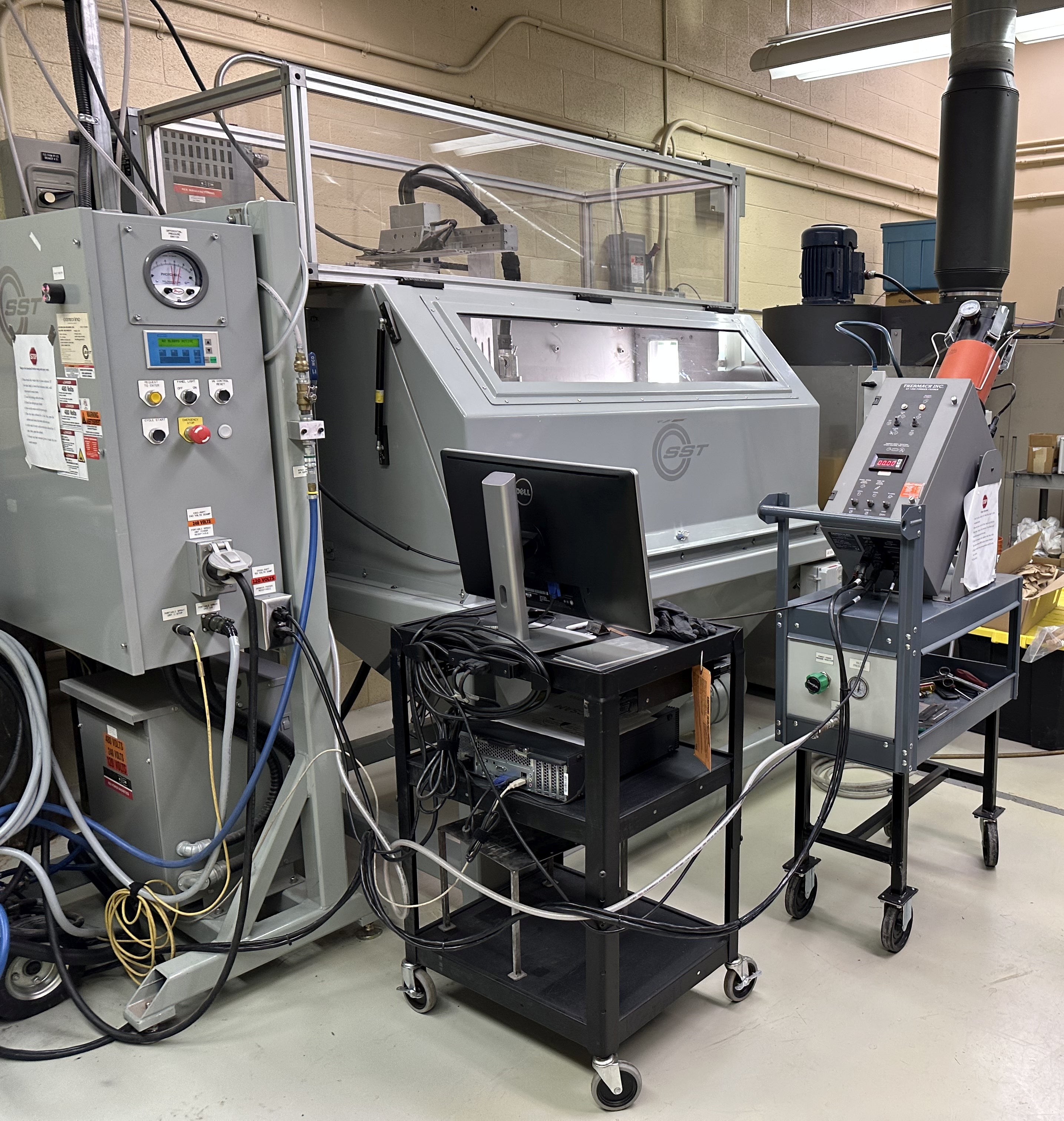 Cold spray setup. SEL controller box on left foreground, PC in center foreground, powder feeder in right middleground, deposition bay in center background.