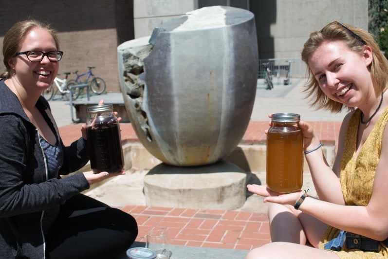 Foregound: Two women sit on a bench holding up large mason jars of tea. Background: Grey fountain of an egg with a large crack