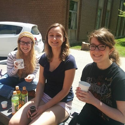 Three woman sit on a bench outside hold various mugs and cups. On the bench there are also three bottles of iced tea.