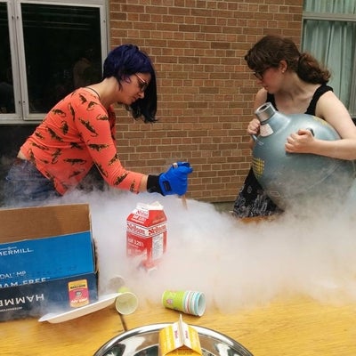 Woman wearing blue rubber gloves stirs. Another woman holds a metal container of liquid nitrogen. Smoke billows around the bowl.