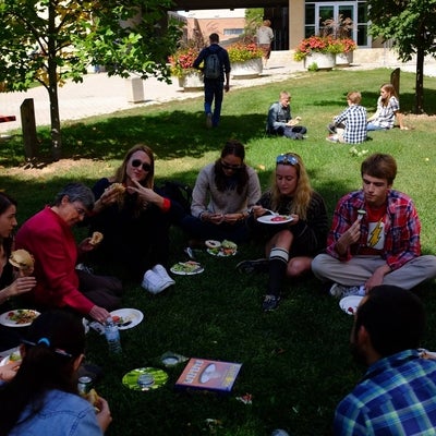 A group of young adults sit on the grass. In front of them is an assortment of food on foam plates.