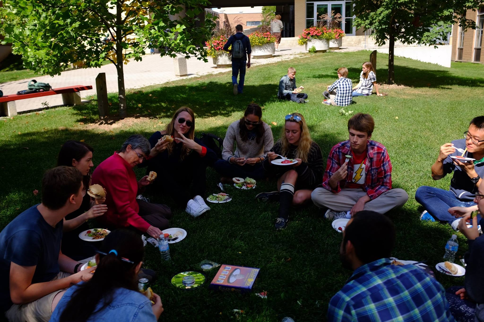 A group of young adults sit on the grass. In front of them is an assortment of food on foam plates.