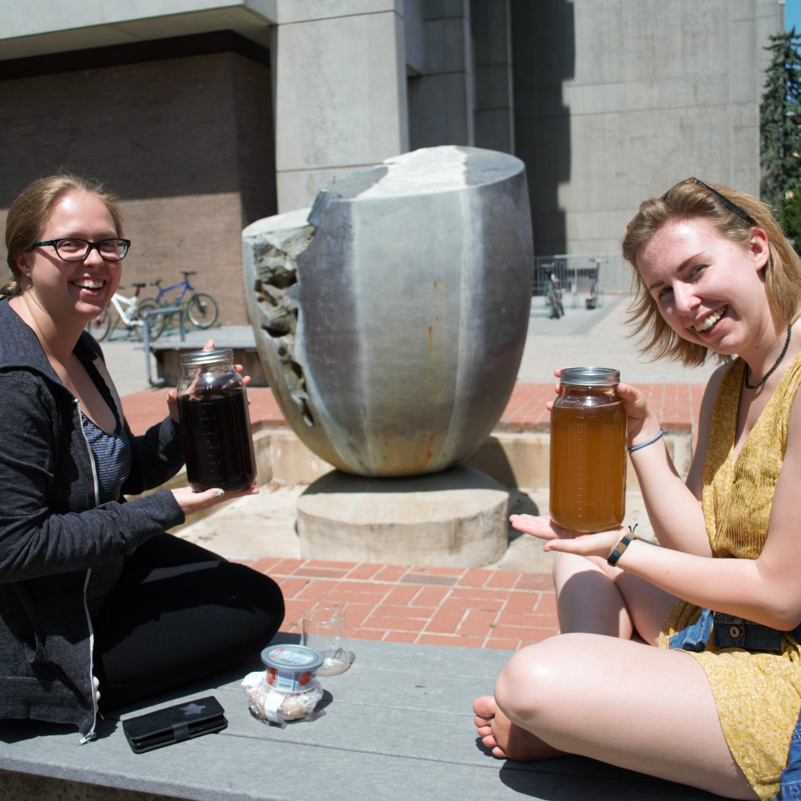 Foregound: Two women sit on a bench holding up large mason jars of tea. Background: Grey fountain of an egg with a large crack