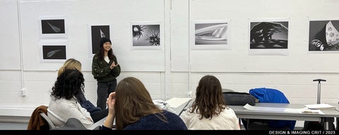 Group of people seated with one standing at the front of the room, in front of several black and white photographs of objects.