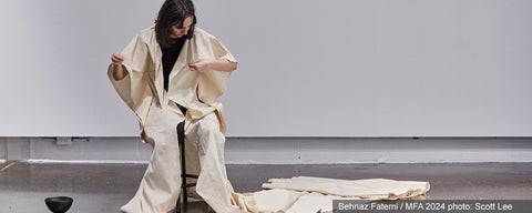 Artistic performance by Behnaz Fahemi. Woman wearing a long canvas robe, sits on a stool and embroiders words in Arabic onto the robe.