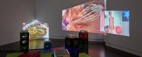 A gallery space with foam floor tiles, vinyl mats and multiple projectors set on stacked plastic crates has overlapping projections of videos on several walls.