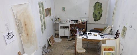 View looking down into a studio with art supplies on a table and shelves and abstract paintings of various sizes on the walls.