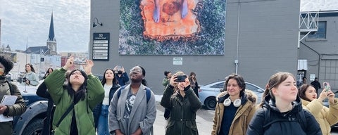 Group of people standing outside, looking up at photographing, and the building behind them has a mural of feet on a tree stump.