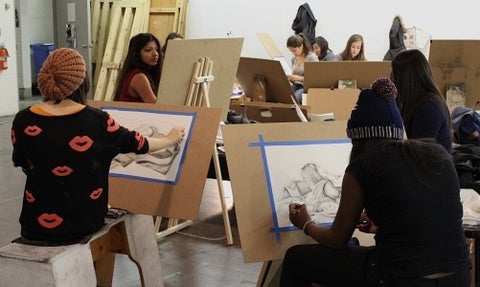 Students in an art class, seated at easels drawing fabric.