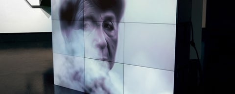 Digital artwork of an elderly woman's face behind smoke, displayed on stacked monitors on the galley floor.