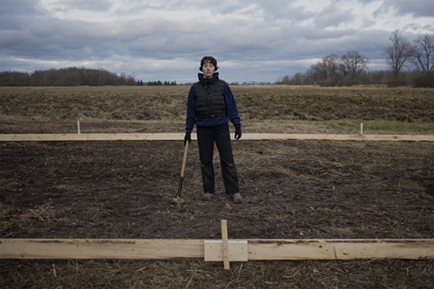 A person holding a long-handled mallet, stands in an area of cleared earth framed by boards, in a farm field. 