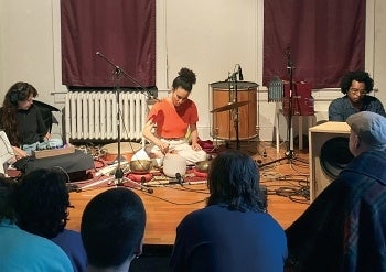 A musical performance with three people sitting on the floor in front of an audience.  The musicians are surrounded by various percussion instruments and electronic equipment.