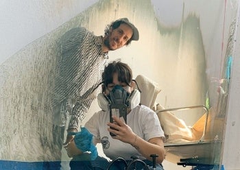 A person wearing a respirator kneels to take a selfie in a reflective surface with a smiling person standing behind them.