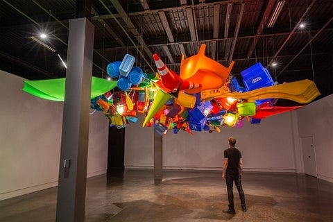 Installation view of a person looking up at a large suspended canopy made up of hundreds of brightly coloured plastic objects.