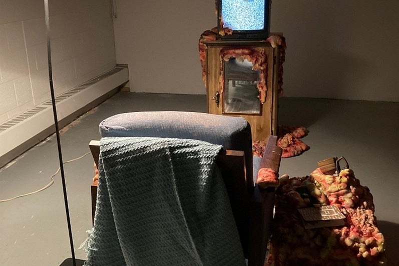 Art installation of an armchair, table, lamp and televison playing static, all topped with spray foam that appears to spread