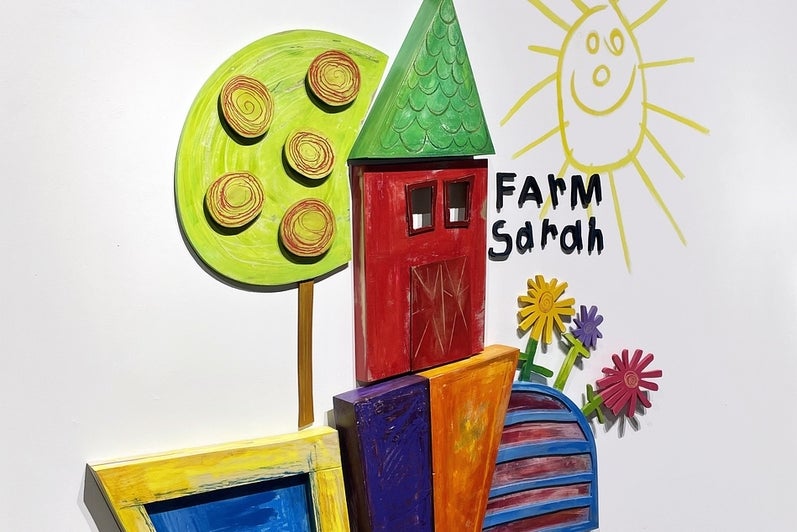 Artwork consisting of painted wooden shapes arranged to form a childlike red barn with a chicken on the roof, tree, and flowers.