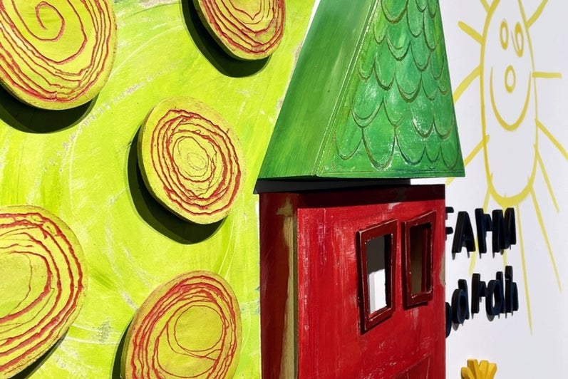 Artwork consisting of painted wooden shapes arranged to form a childlike red barn with a chicken on the roof and tree