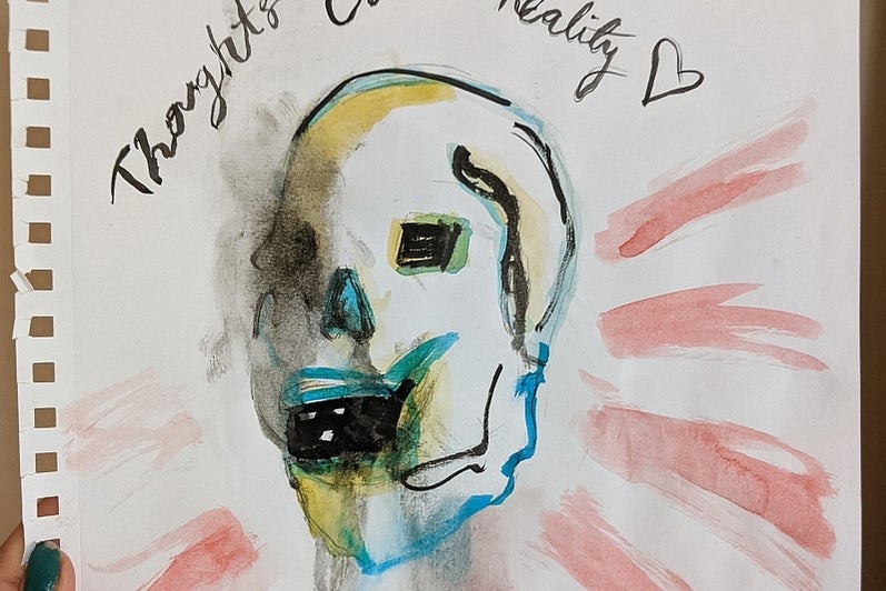 Hand holds a watercolour painting of a skull with the text "thoughts create reality"