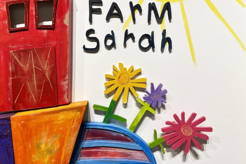 Detail of artwork consisting of painted wooden shapes arranged to form a childlike red barn and flowers, text "Farm Sarah"