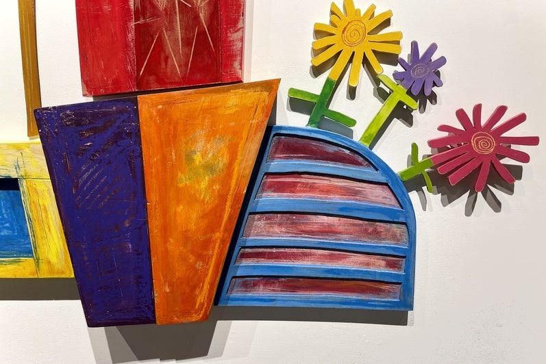 Detail of artwork consisting of painted wooden shapes arranged to form a childlike red barn and flowers