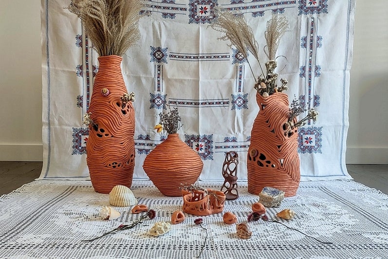 Collection of coiled clay vessels with dried flowers and shells sit on the floor on a crocheted table cloth with an embroidered