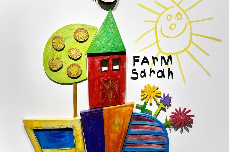 Artwork consisting of painted wooden shapes arranged to form a childlike red barn with a chicken on the roof, tree, and flowers.