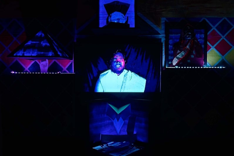 Art installation of monitor and lit scultpures arranged in a cross pattern.  Central monitor show a man against a black backgrou