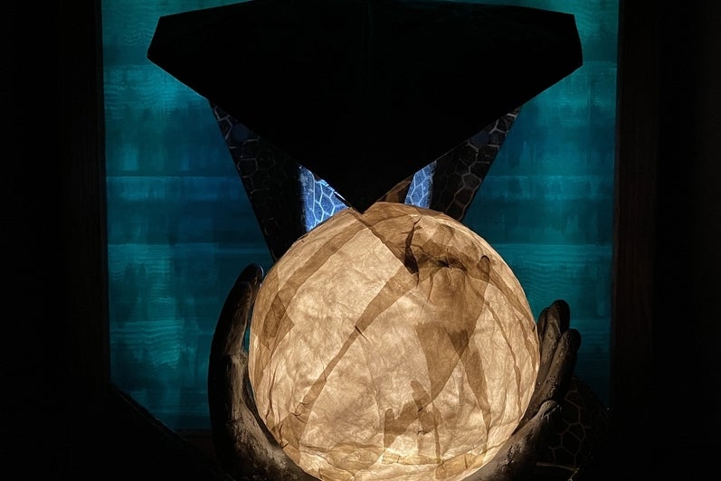 Artwork of glowing papier mache sphere is held in two hands with a paper cobra head above against a pattern teal coloured backgr
