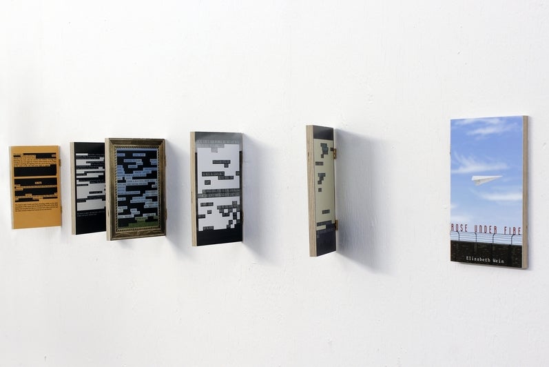 Art installation of 6 book covers attached to the wall with hinges, 5 are opened to reveal the back text with redacted words.