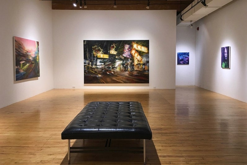 View of an art gallery with an exhibition of paintings.  The painting directly in front show a Hong Kong street scene at night.