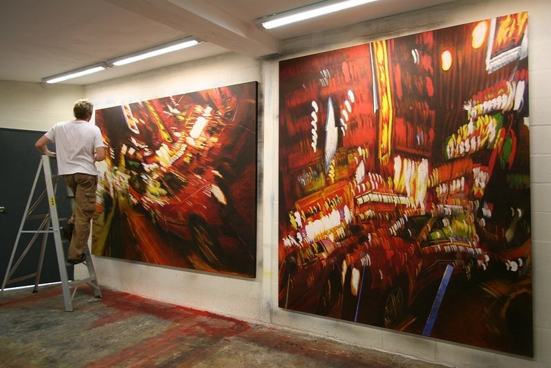 Artist on a step ladder at work in a studio on two, predominantly red and white, paintings of neon street signs at night viewed with a slight motion blur.