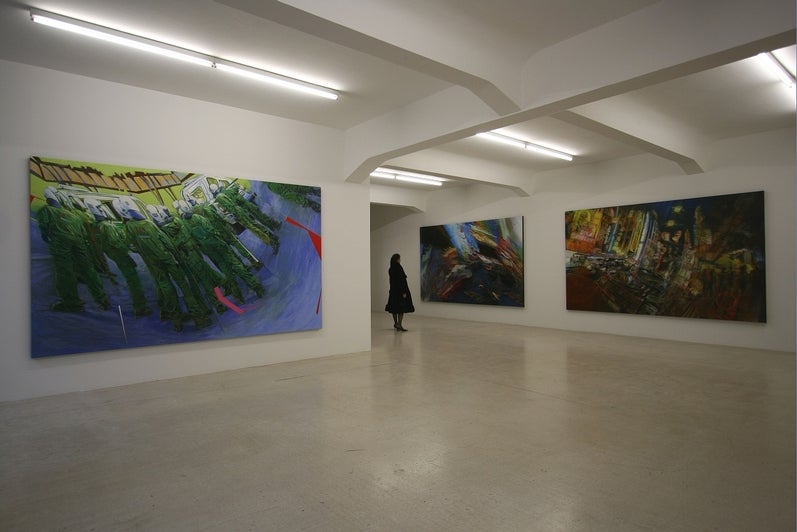 View of an art gallery with a person viewing the exhibition of paintings.  The two paintings on the right are large colourful street scenes with a blur of street signs at night; the painting on the left shows a line of police in riot gear with anamorphic distortion.