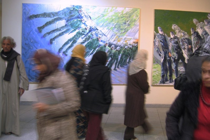 Motion blur showning multiple people, several of them wearing head scarves, moving in front of two paintings each depicting a line of police in riot gear with anamorphic distortion. 