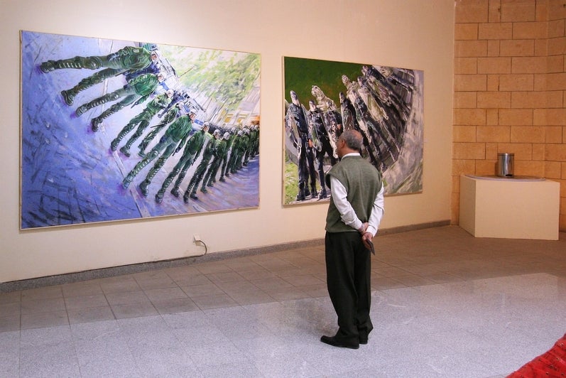 A man stands in front of two paintings each depicting a line of police in riot gear with anamorphic distortion.