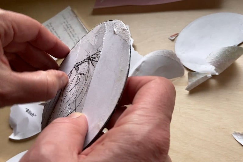 Two hands peal back paper to reveal a pen and ink drawing of a tree