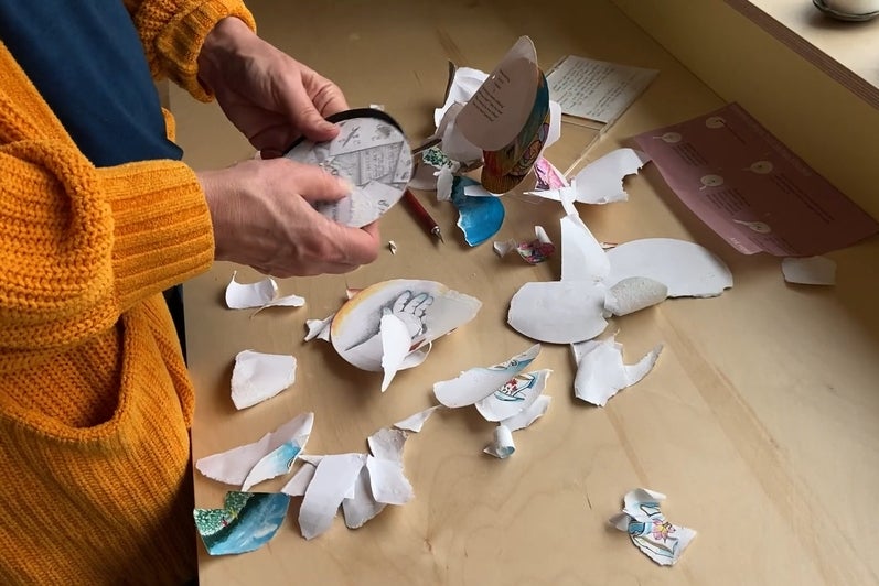 Person holding a circular drawing stands by a table covered with bits of torn drawings.