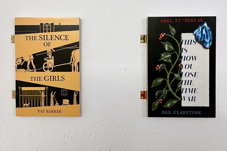 Art installation of 2 book covers attached to the wall with hinges titled "The silence of the girls" and "This is how you lose t