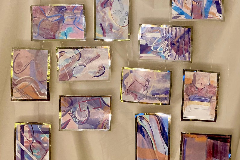 Twelve small abstract paintings in shades of mauve, blue, brown and gold hang suspended against a cloth.