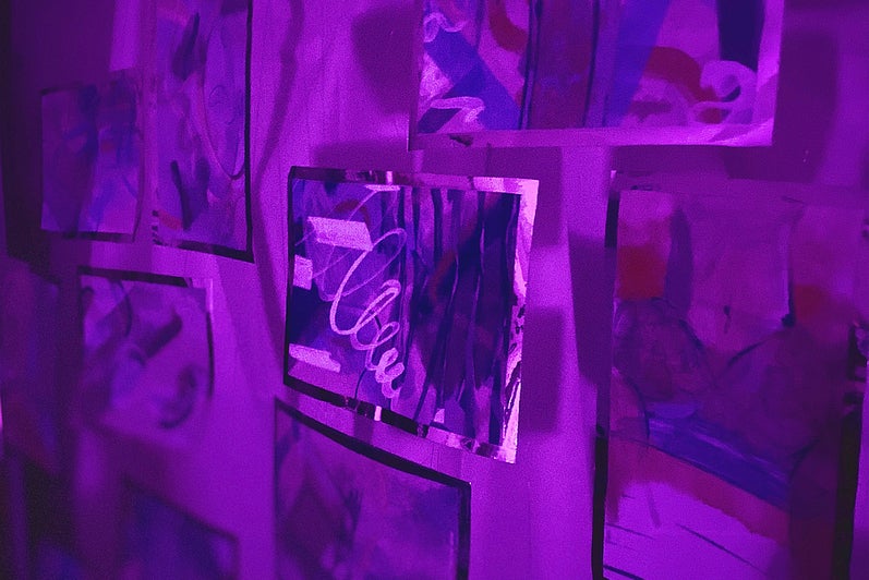 Deep purple light on10 small abstract paintings hanging suspended.  Artworks edged with reflective material.