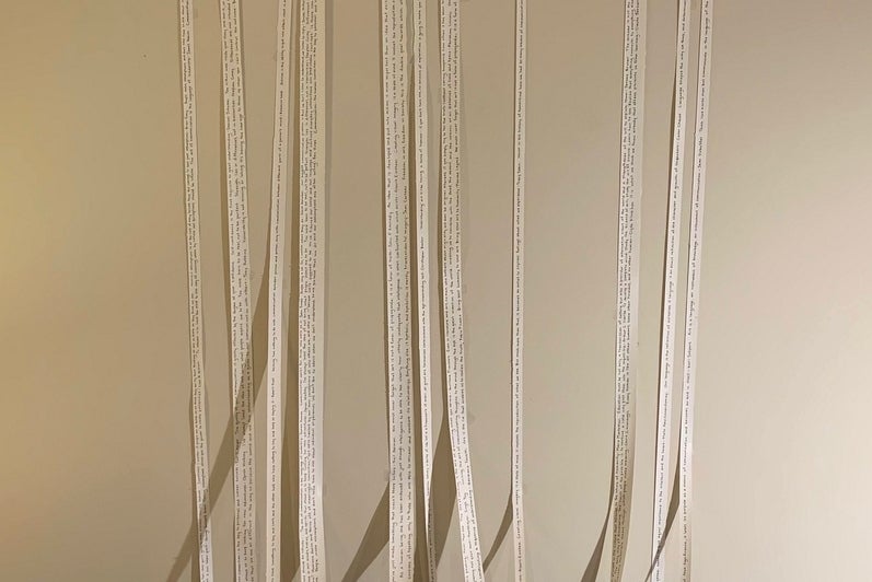 Art installation of three books lying on the floor with strips of paper coming from inside the books and stretched up the wall