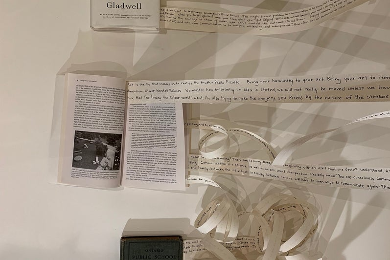 Art installation of three books lying on the floor with strips of paper coming from inside the books and stretched up the wall